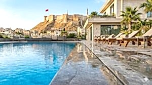 Gündüz çekilmiş, berrak suyuyla parlayan modern bir taşmalı havuzun kenardan görünümü. Sağ tarafta lüks bir villa ve güneşlenme şezlongları, arka planda ise tarihi Gaziantep Kalesi yükseliyor.